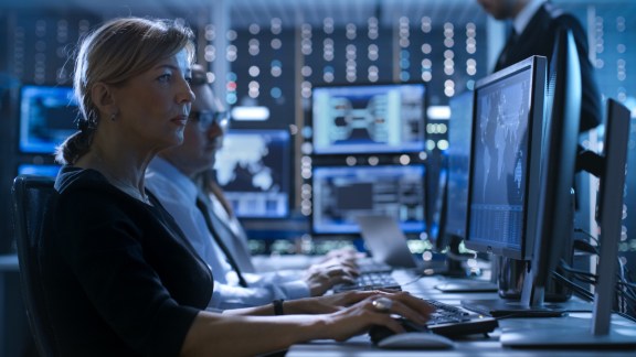 Female Government Employee Works in a Monitoring Room. In The Background Supervisor Holds Briefing. Possibly Government Agency Conducts Investigation.