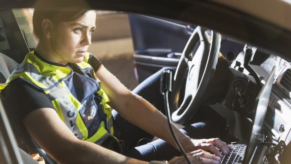 A female police officer sitting in her police car, using the computer. It is nighttime and the emergency lights on her vehicle are flashing.