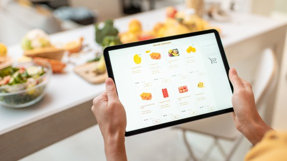 Woman shopping food online using a digital tablet at the kitchen, close-up view on a tablet screen. Concept of buying online using mobile devices