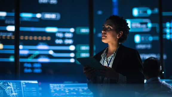 Young Multiethnic Female Government Employee Uses Tablet Computer in System Control Monitoring Center. In the Background Her Coworkers at Their Workspaces with Many Displays Showing Technical Data.