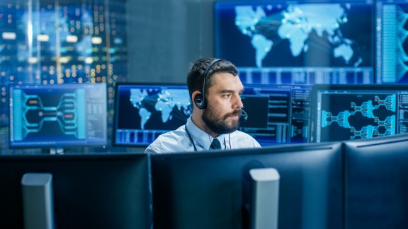 System Control Room Dispatcher Talks into Headset. He Controls Correct Work of the Facility. In the Background Multiple Monitors Show Technical Data.