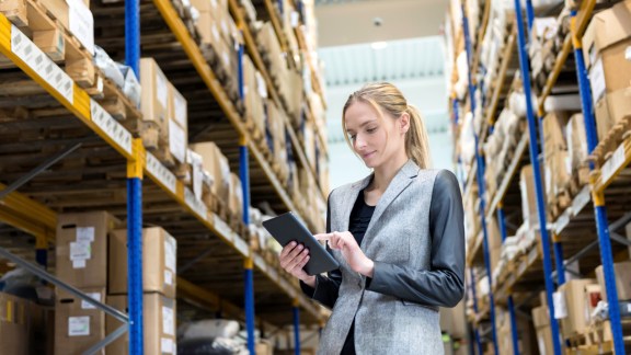 Portrait of a pensive young woman supervisor working on-line in warehouse. Young blond woman standing at distribution warehouse and wearing elegant suit. Industrial boss examining the stock. Large distribution storage in background with racks full of packages, boxes, pallets, crates ready to be delivered. Logistics, freight, shipping, receiving.