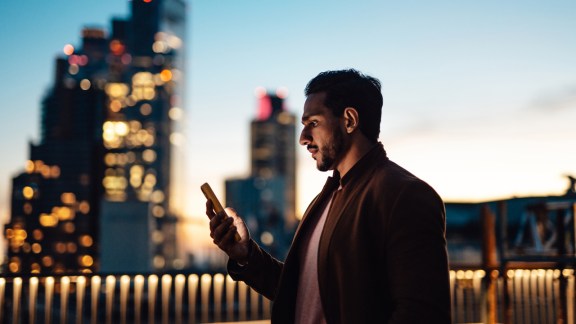 Businessman using mobile phone at rooftop in business building against illuminated financial buildings and London cityscape at night.