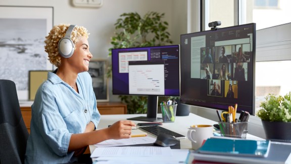 Cheerful businesswoman holding document during video conference with colleagues. Smiling blond Afro female professional is working at home office. She is wearing wireless headphones.