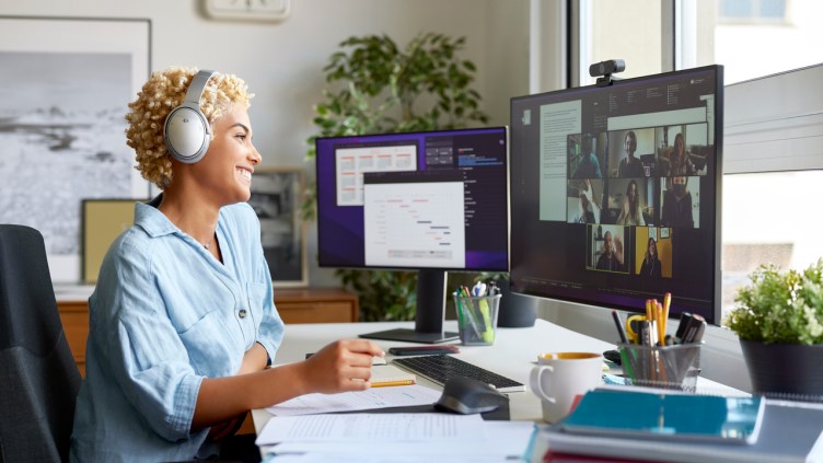 Cheerful businesswoman holding document during video conference with colleagues. Smiling blond Afro female professional is working at home office. She is wearing wireless headphones.