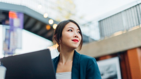 Low angle view of young Asian business woman looking away while working on laptop, sitting at sidewalk cafe. Remote working concept. Freelancer working from cafe.