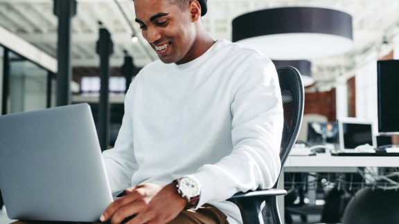 Smiling businessman working on a laptop in a modern office. Happy young businessman enjoying working alone in a modern workplace. Creative young businessman working on a new project.