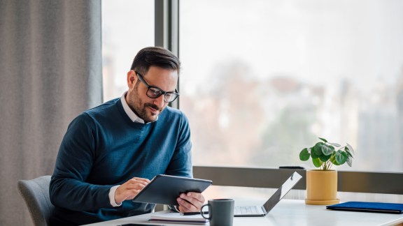 Young businessman using digital tablet while working on laptop in business office. Male professional with wireless computer at desk. He is sitting by window in office.