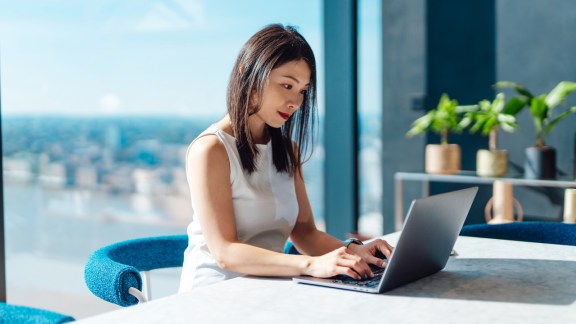 Young Asian woman working with laptop while sitting by the window with city view and sunlight.