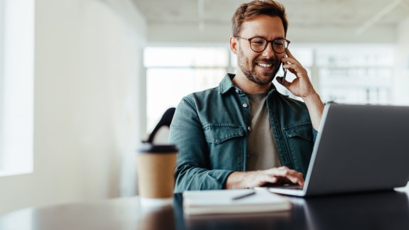 Software designer discussing a new project with his client on the phone. Creative business man working on a laptop in an open plan office.