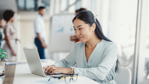 Asian woman working on laptop. Businesswoman busy working on laptop computer at office with colleagues in the background.