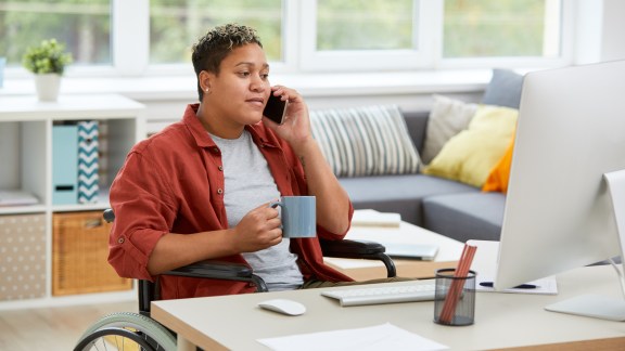 African disabled woman sitting in wheelchair at the table and looking at computer monitor while talking on mobile phone and drinking coffee at home