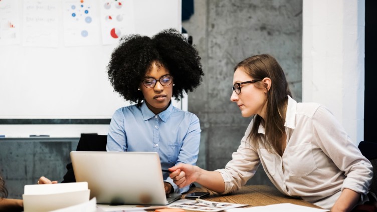 A black and a caucasian young woman are sitting in a business meeting in a modern office.They discuss something over papers and a laptop while one of them is pointing at the computer screen. Both are wearing glasses and pie charts are visible in the background.