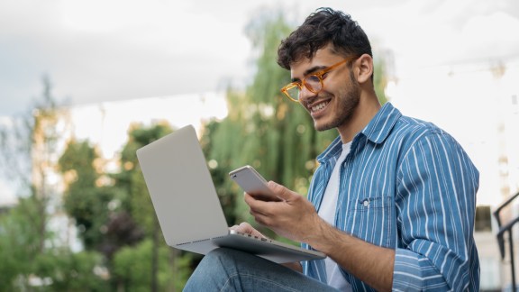 Young handsome Indian man using laptop computer, mobile phone, working freelance project online, sitting outdoors. Successful business. Asian student studying, learning language, online education concept