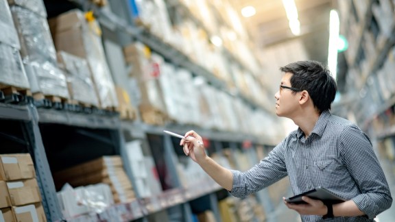 Young Asian man worker doing stocktaking of product in cardboard box on shelves in warehouse by using digital tablet and pen. Physical inventory count concept