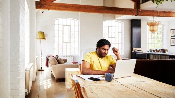 A guy wearing a yellow t-shirt works from home using his dining room table as a desk