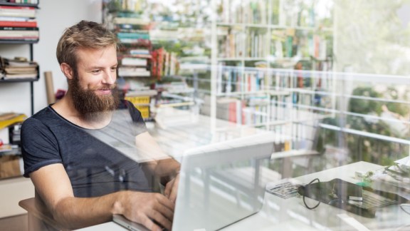 Young bearded man sitting on desk in his home office and working on laptop.