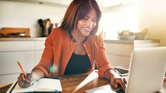 Smiling young African female entrepreneur writing down notes and working on a laptop while sitting at her kitchen table