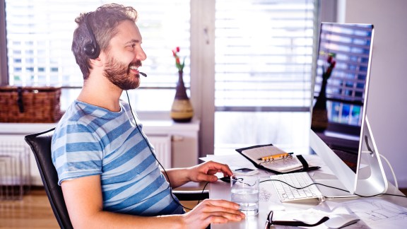 Man sitting at the desk working from home on computer, talking on the phone, headset on head, smiling