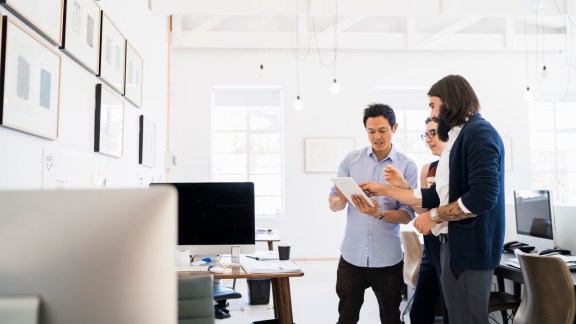 A photo of creative business people using digital tablet. Colleagues standing while holding technology at workplace. Professionals are working in office.