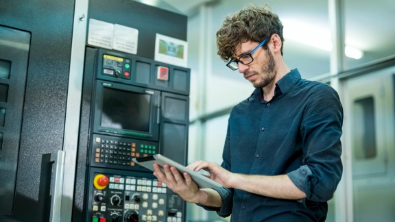 Expert of Male Software engineer working in front of automated CNC machine. He is analyzing machine operation problems by digital tablet.