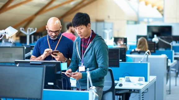 Two men using mobile phones at desk with computers in modern open space of large tech company.